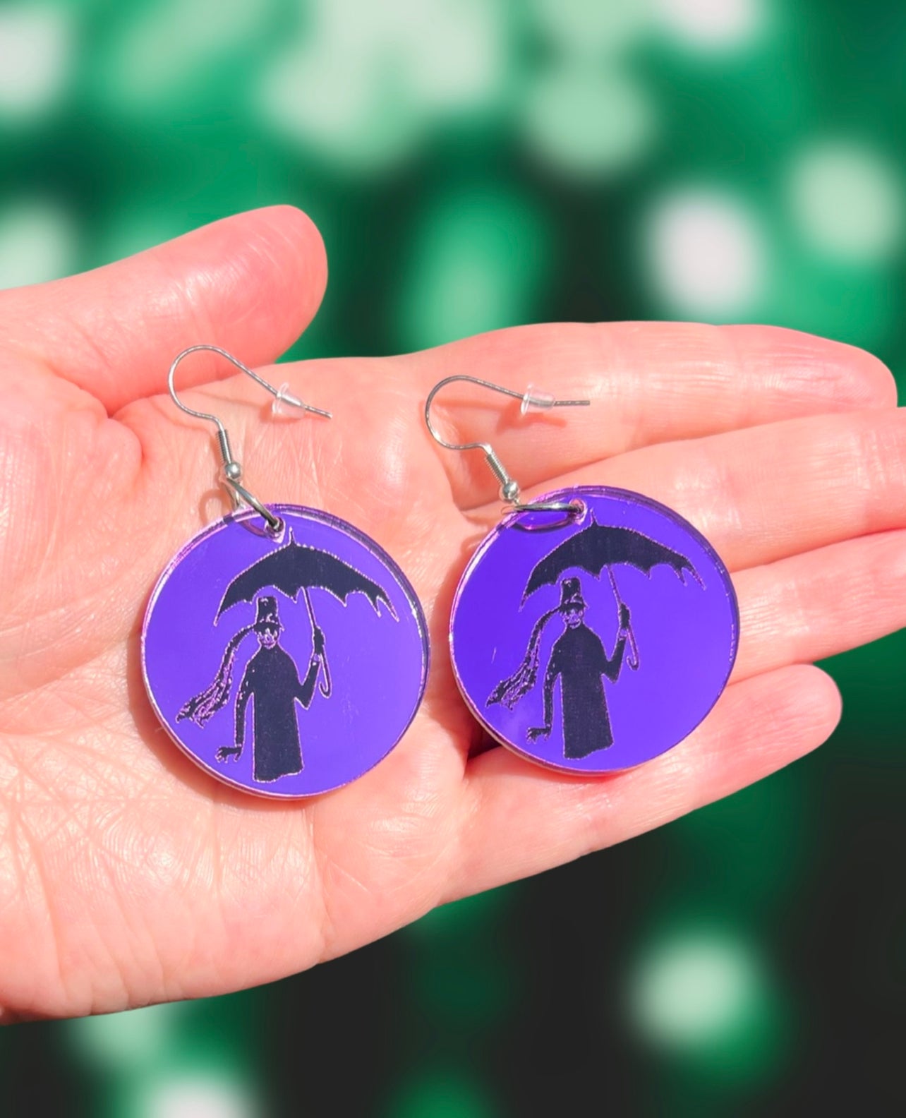 small round purple earrings, with the image of a dark figure holding an umbrella engraved on them. They are adorned with hooks and are resting on an open hand in front of a green blurry background.