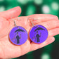 small round purple earrings, with the image of a dark figure holding an umbrella engraved on them. They are adorned with hooks and are resting on an open hand in front of a green blurry background. 